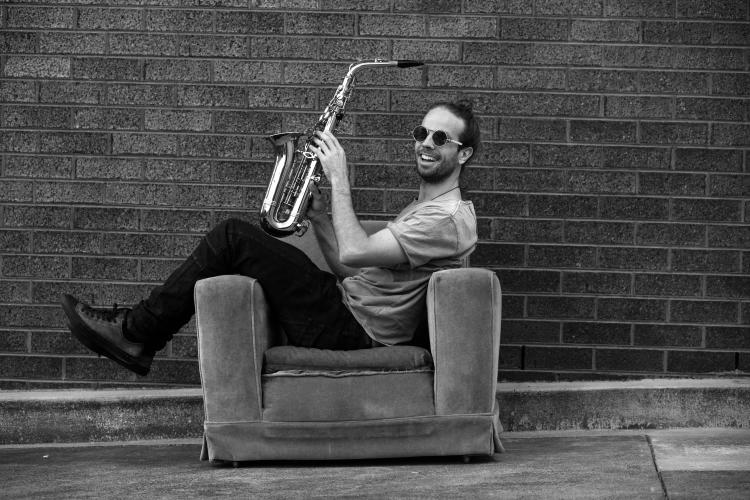 Cheshire sitting sideways on a couch holding a saxophone against a brick wall