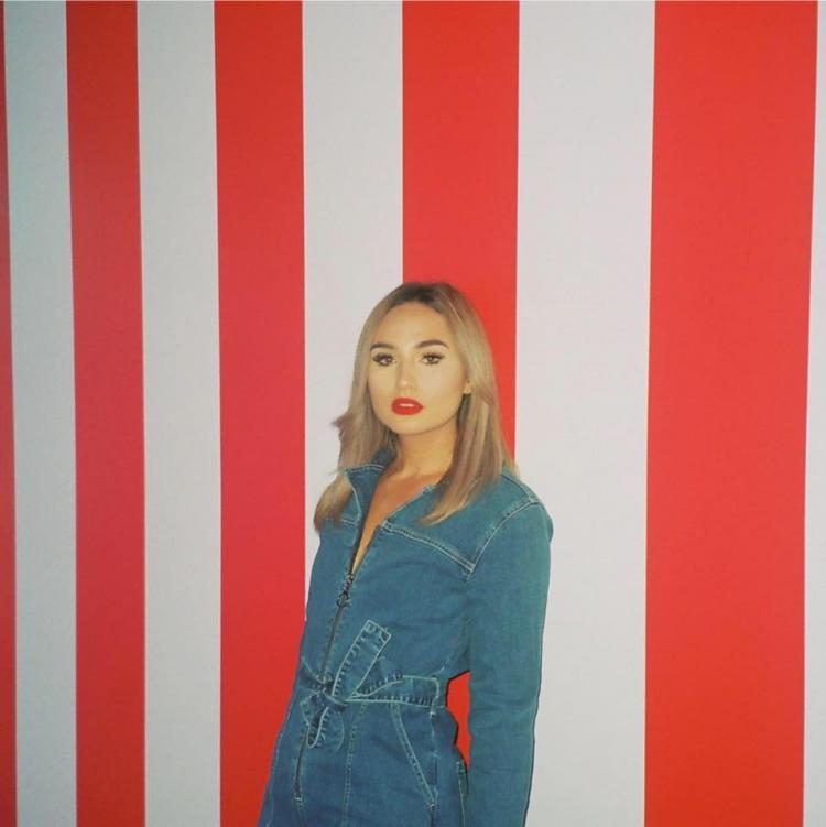 DVNA in a denim jumpsuit against a red and white background.