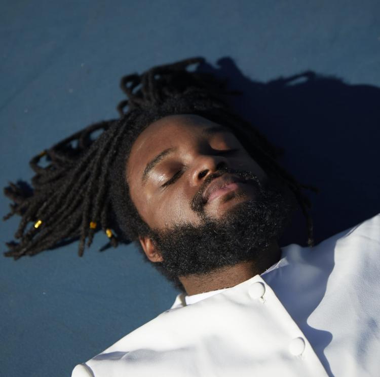 Genesis Owusu in a white shirt laying day with eyes closed on a blue background