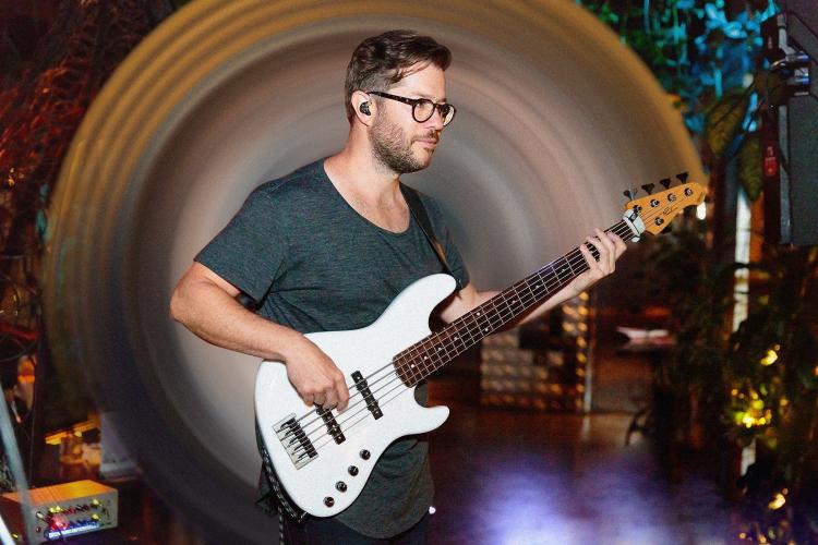 Mark Peric holding a white bass guitar with a dragged effect from the headstock around Marks head. Black background.