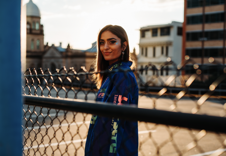 SKRAM standing behind a fence in the broad daylight wearing a blue jacket with colorful sleeves