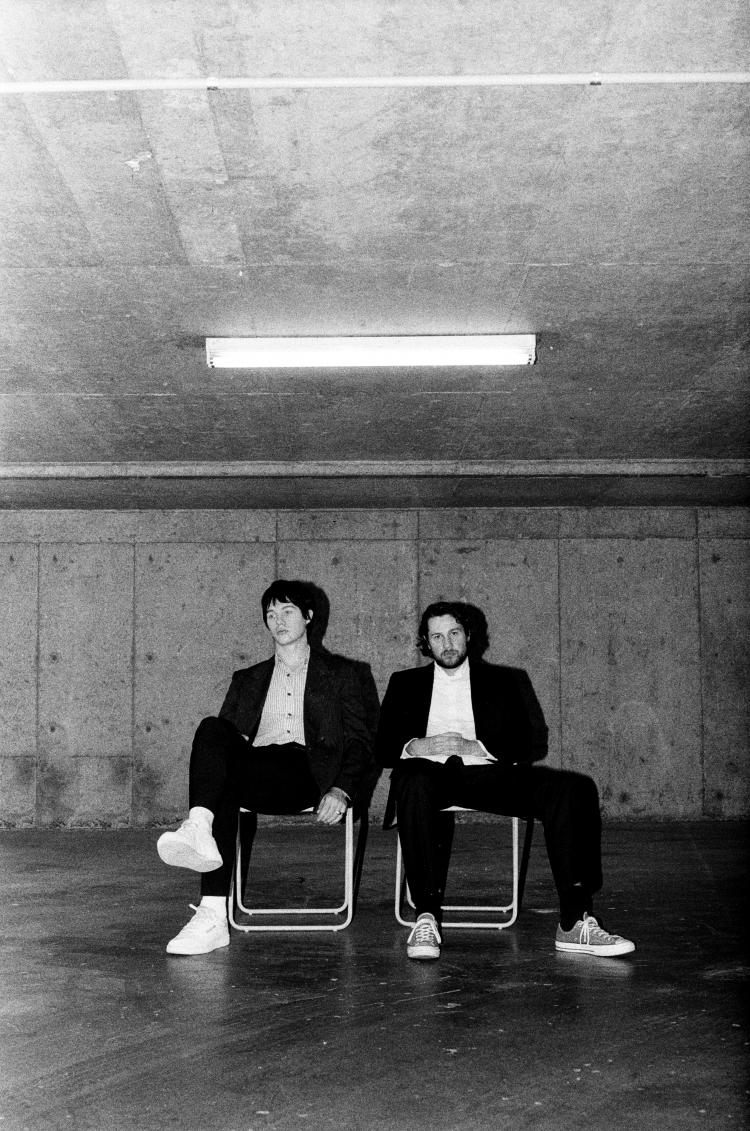 SUPERNEW in a black and white image sitting on deck chairs under a fluorescent light in an empty garage looking room.
