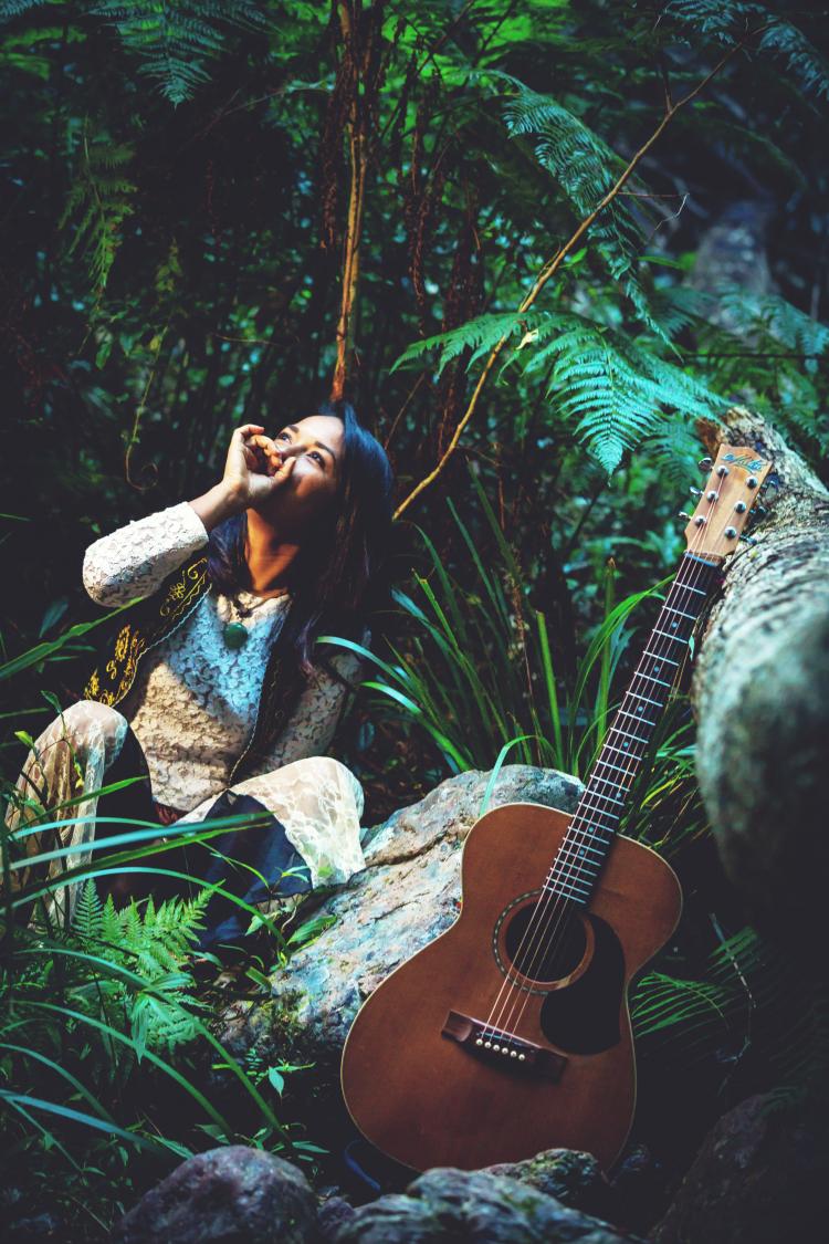 Alisha Todd sitting in a forest holding her mouth with her guitar in the forefront of the photo.