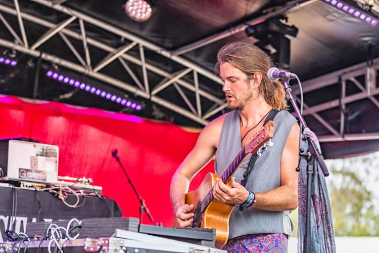 B-Syde playing his guitar live in a grey vest with a DJ deck on the right hand side of the image in front of a red background.