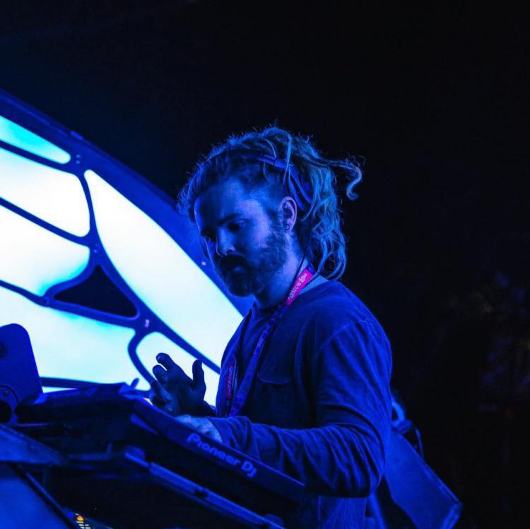 BustaFlux in a blue lit room DJing facing to the left of the frame. He has dreadlocks and has a red lanyard around his neck. There are lights behind him.