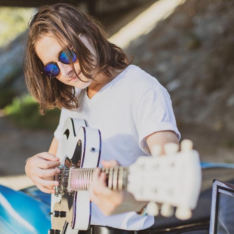 Harry J Hart in a white shirt and sunglasses holding a white guitar with the headstock aimed at the camera.