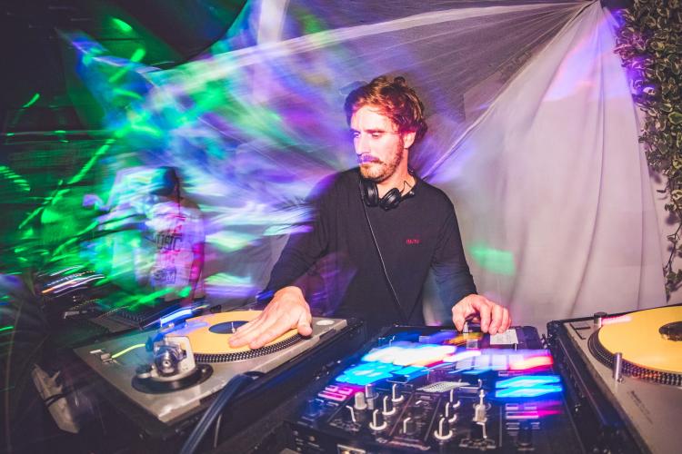 Mr. HA YES DJing in a psychedelic setting wearing a black shirt with headphones around his neck.