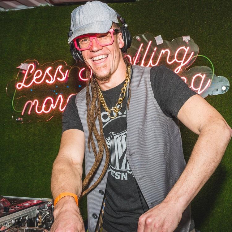 ZygoTwitch DJing in a blue hat, red glasses, a black t-shirt and a grey vest against a black background.