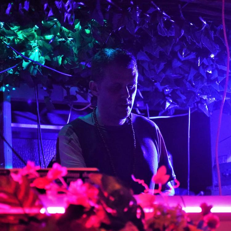 pH-Zero DJing in a low lit room with bright pink, green and blue lights. Wearing a necklace and black t-shirt.